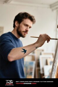 Medium shot of a man's forearm with a bird tattoo, holding a paintbrush in a navy sweater.