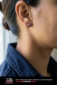 Dainty dotwork crescent moon tattoo behind ear on deep brown skin.