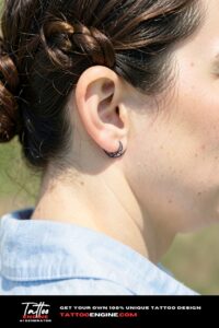 Dainty geometric crescent moon tattoo behind ear on beige skin, bright afternoon light.