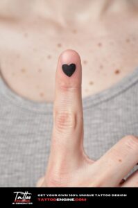 Tiny bold black heart tattoo on middle fingertip, closely cropped on pale, freckled skin.