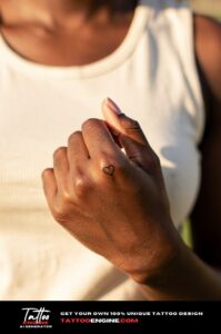 Tiny heart outline tattoo on underside of pinky finger, fine line on dark brown skin.