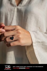 Small solid black heart tattoo on the side of a pinky finger, healed on tan skin.
